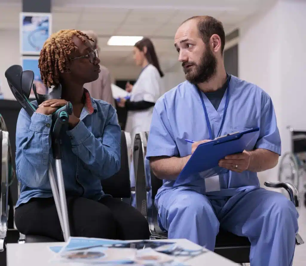 Medical asisstant consulting woman with crutches in hospital reception lobby, talking about diagnosis and medicine in waiting room area. Diverse people discussing at checkup appointment.