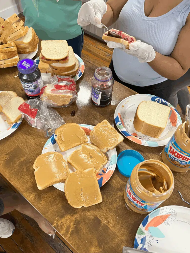 Volunteers wearing gloves preparing peanut butter and jelly sandwiches on a wooden table.