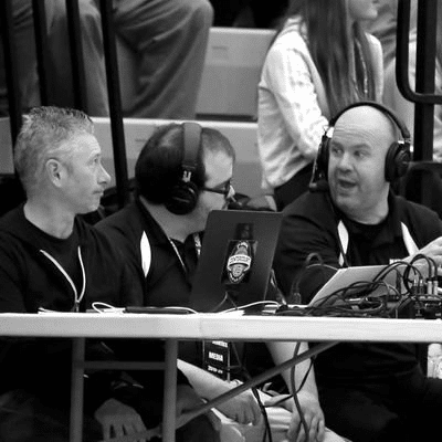 Three commentators with headphones and audio equipment sitting at a table in front of bleachers.