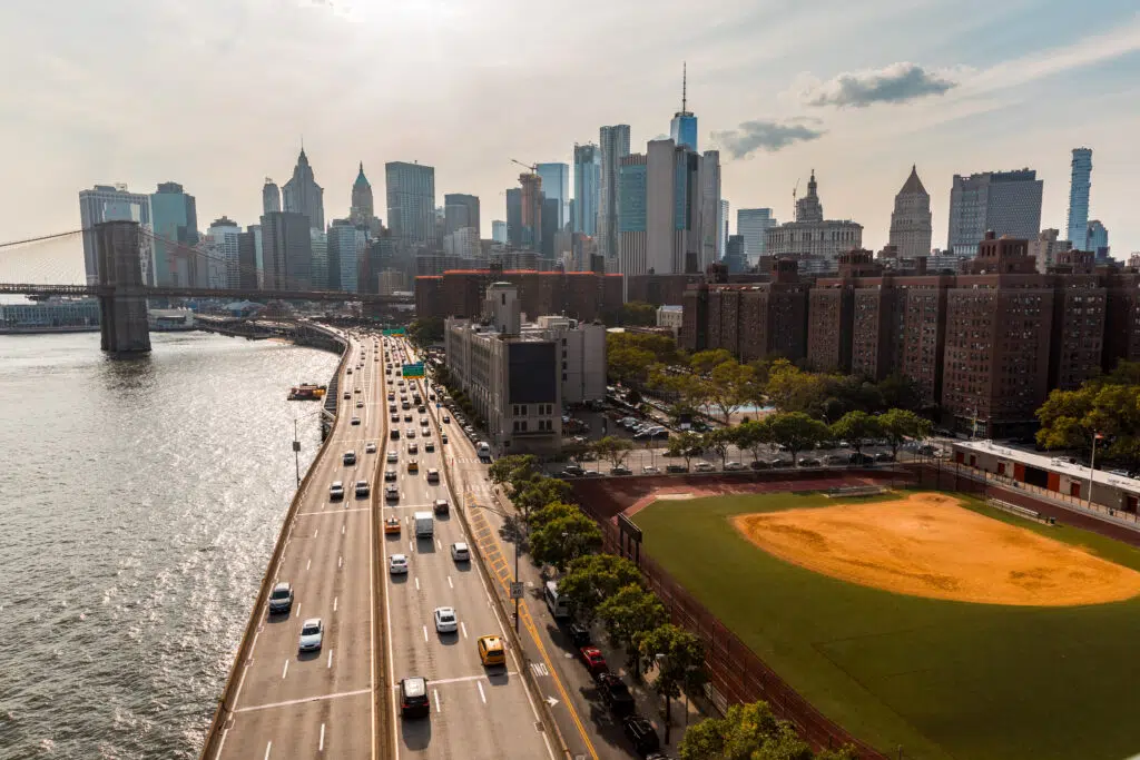 This is an aerial shot of a large highway with many vehicles traveling on it. In the background is a dense cityscape of tall buildings and skyscrapers. To the side of the highway is a baseball field and trees.