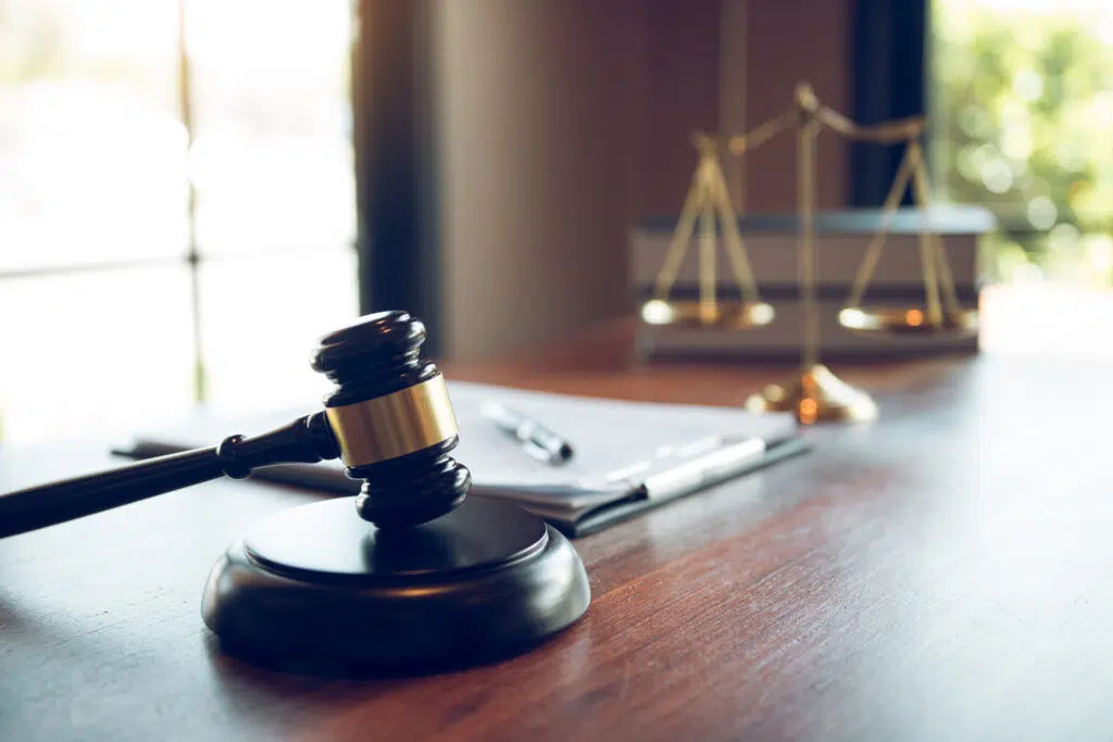 A gavel, scales of justice, pen, and paperwork rest on a wooden desk in this indoor shot. The scales of justice are slightly out of focus in the background.