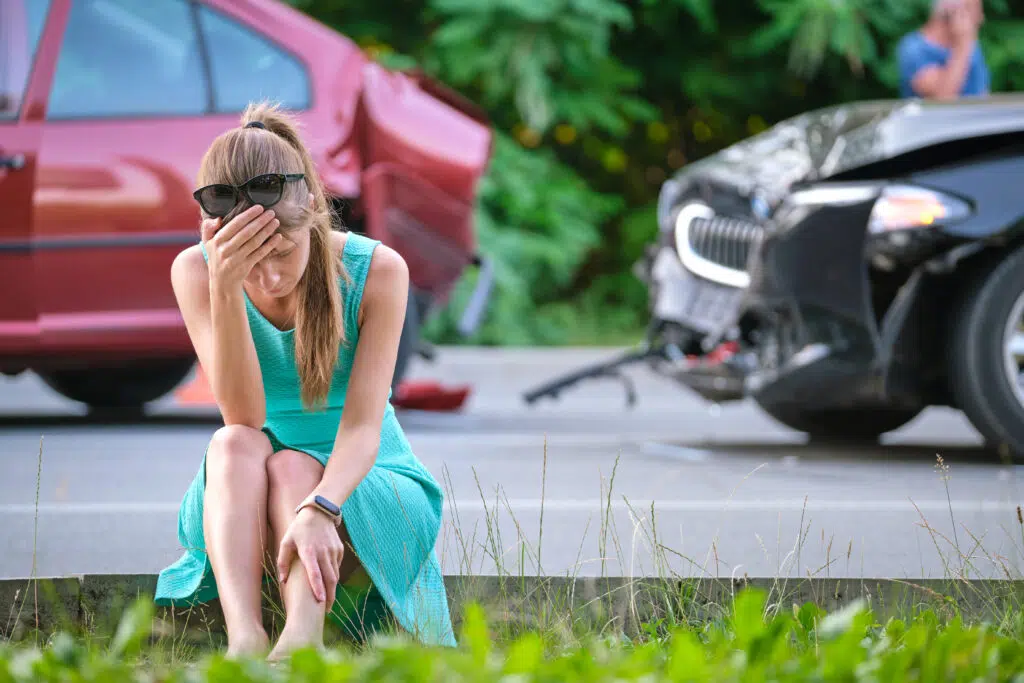 Sad female driver sitting on street side shocked after car accident. Road safety and vehicle insurance concept.