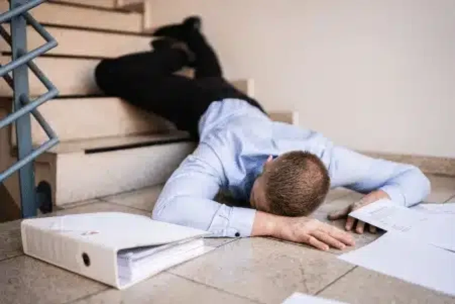 A man in business attire lies face-down on a stairway landing after an apparent fall, with scattered papers and a binder around him